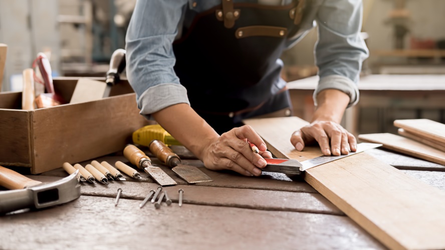 Ein Handwerker zeichnet mit einem Winkel etwas auf einem Brett an, daneben liegen Handwerkzeuge wie Beitel, Sägen, ein Hobel